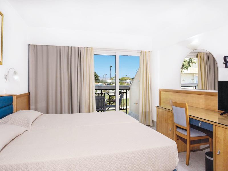 Světý hotelový pokoj s velkou postelí, psacím stolem a balkonem s výhledem na bazén.