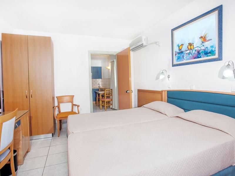 Světý hotelový pokoj s manželskou postelí, skříní, židlí, stolem a televizí.