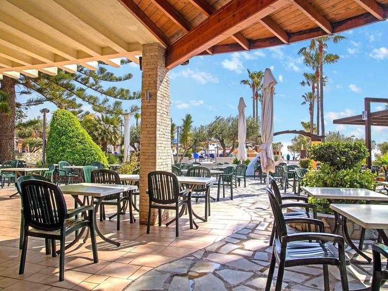 Zastřešená terasa s stoly a židlemi s výhledem na slunečníky a rostliny v venkovní části hotelu.