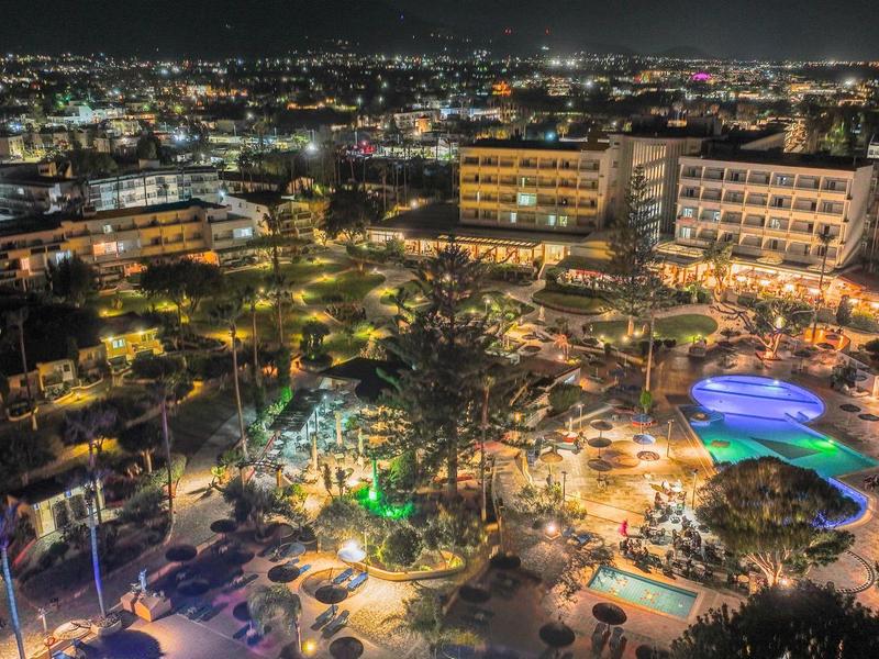 Noční pohled na jasně osvětlený bazén resortu s okolními budovami a městskými světly v pozadí.