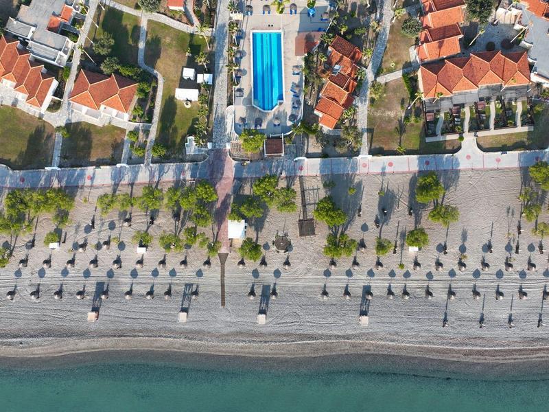 Pohled z ptačí perspektivy na pláž s uspořádanými slunečníky, promenádou a domy v pozadí.