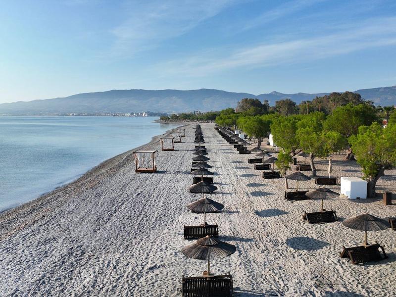 Rozsáhlá oblázková pláž se slunečníky a lehátky vedle klidného moře a hor.