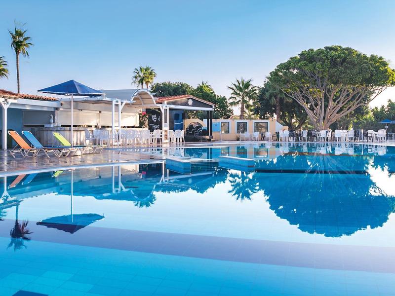 Velký bazén s modrou vodní hladinou před bungalovy a palmami pod jasnou oblohou.