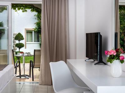 Licht hotelkamer met bureau, tv en uitzicht op een terras met tuinmeubilair.