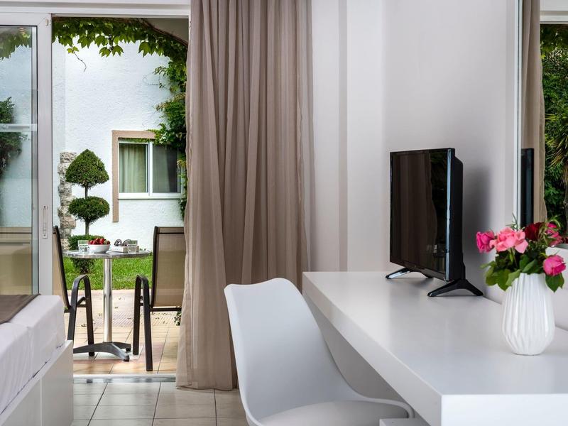 Licht hotelkamer met bureau, tv en uitzicht op een terras met tuinmeubilair.