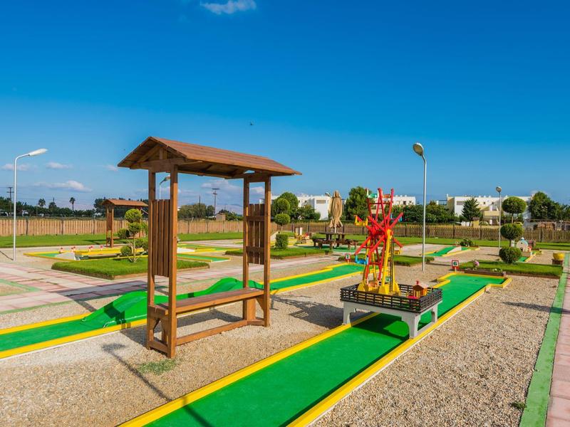 Buitenspeelplaats voor minigolf met groene banen en houten bankjes, omringd door gras en gebouwen.