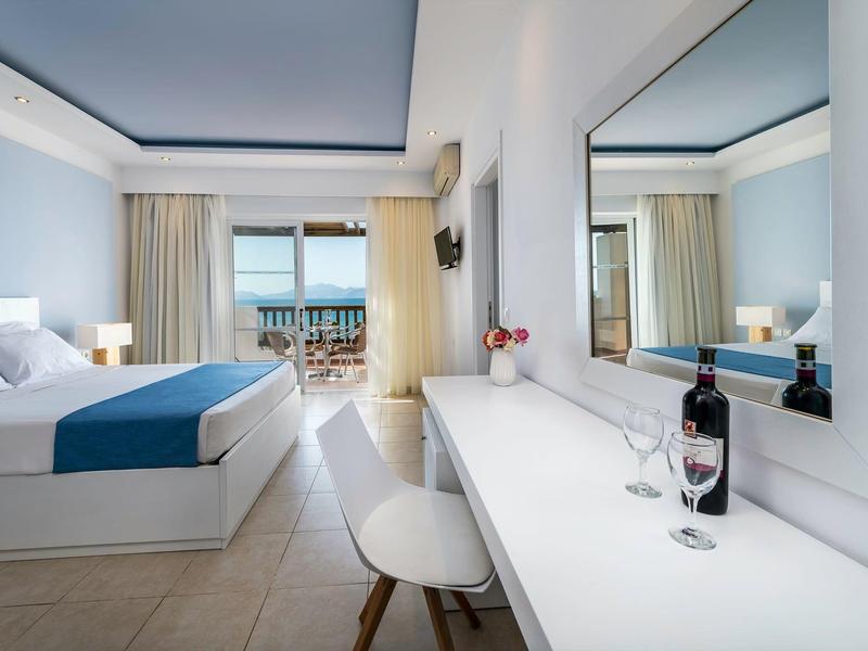 Lichte hotelkamer met bed, bureau, stoelen en balkon met uitzicht op zee.