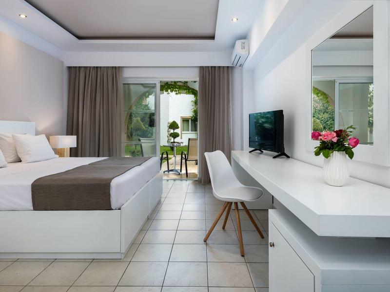 Lichte hotelkamer met tweepersoonsbed, bureau, stoel en uitzicht op een balkon met tuin.