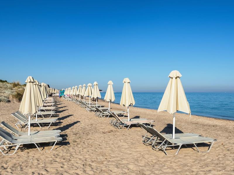 Lege parasols en ligstoelen in rijen op een zandstrand bij helderblauwe lucht.