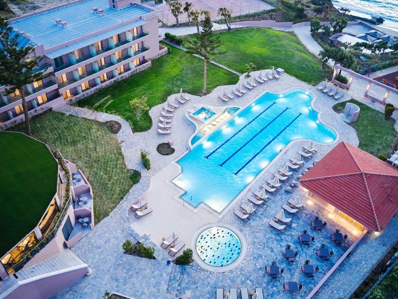 Verlicht hotelzwembad met ligstoelen en jacuzzi in een tuin bij schemering.