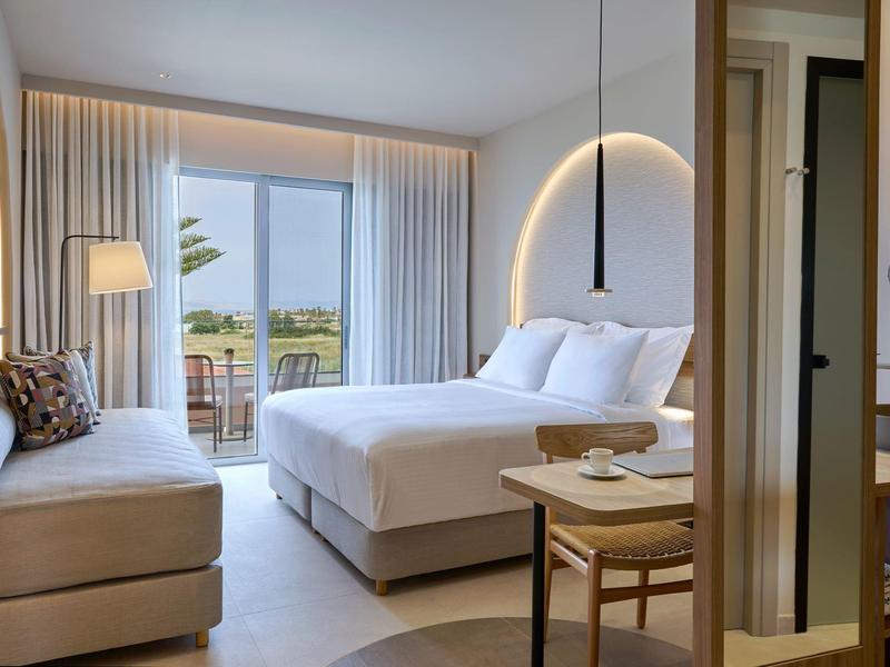 Moderne hotelkamer met tweepersoonsbed, bank en balkon met uitzicht op palmbomen.