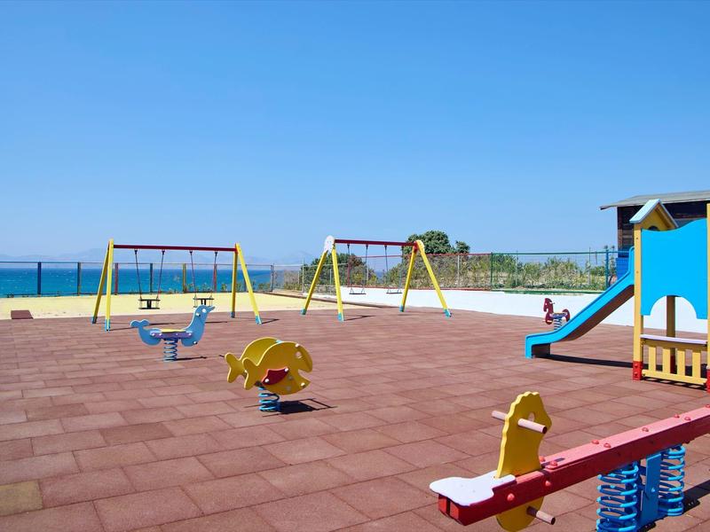 Speeltuin aan zee met kleurrijke schommels, glijbaan en wip onder een heldere hemel.