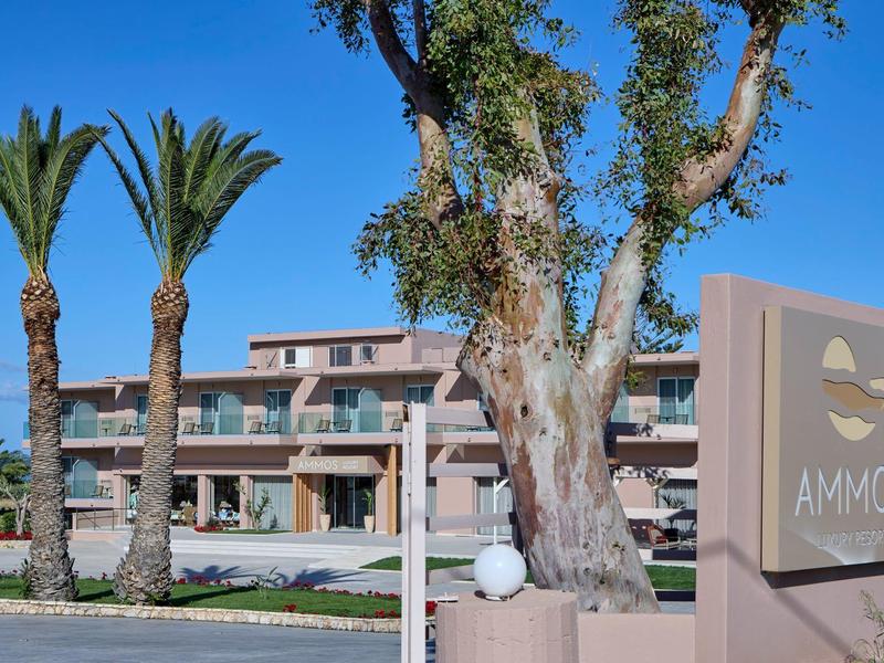 Modern hotelcomplex met grote bomen en palmbomen onder een heldere blauwe lucht.