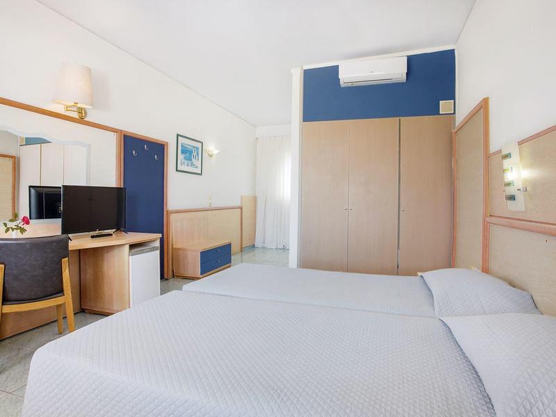 Jasny pokój hotelowy z dwoma łóżkami, drewnianymi meblami, biurkiem i telewizorem na białej ścianie.