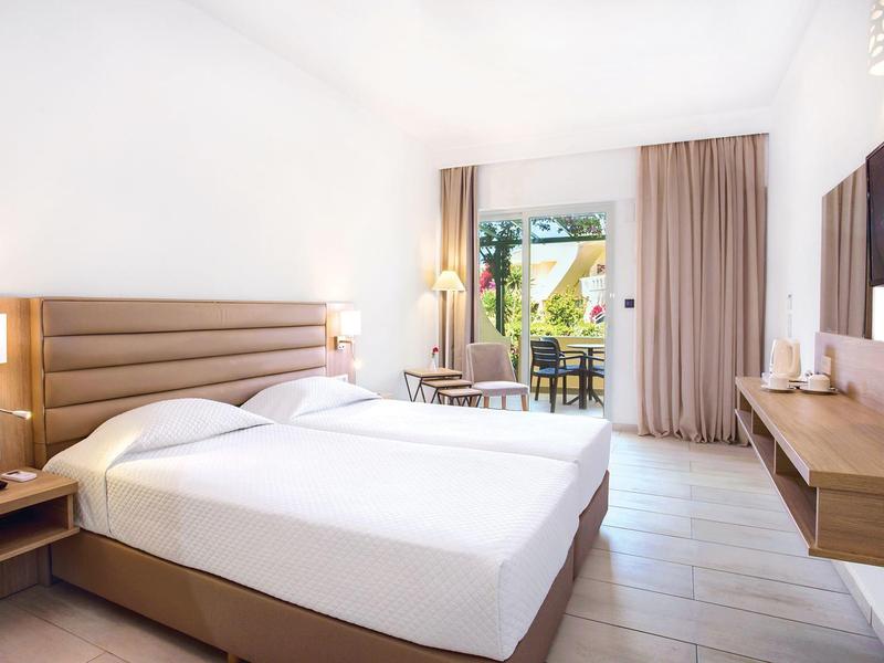 Jasny pokój hotelowy z dużym łóżkiem, drewnianymi meblami i widokiem na balkon.