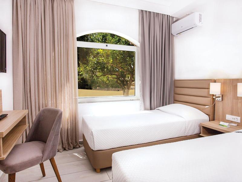 Jasny pokój hotelowy z dwoma łóżkami, krzesłem, biurkiem i oknem z widokiem na zieleń.