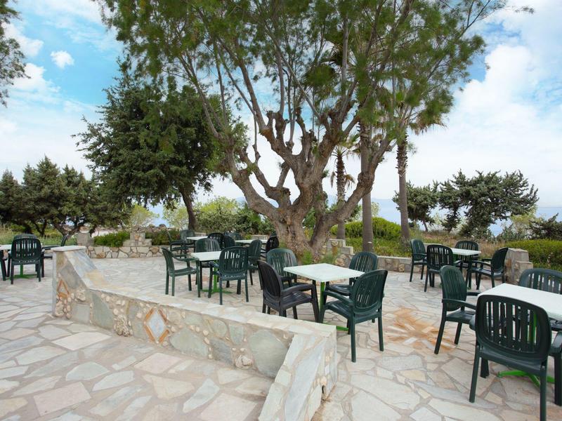 Zewnętrzny taras z zielonymi krzesłami i stołami na kamiennej podłodze pod drzewami w słoneczny dzień.