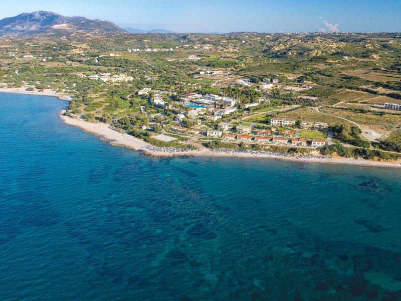 Krajobraz nadmorski z czystą niebieską wodą, piaszczystą plażą i rozproszonymi budynkami przy zielonych wzgórzach.