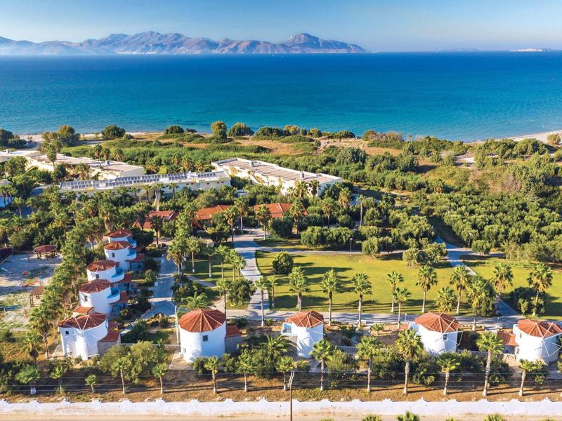 Nadmorski kurort z białymi bungalowami, bujną zielenią i widokiem na błękitne morze oraz odległe góry.