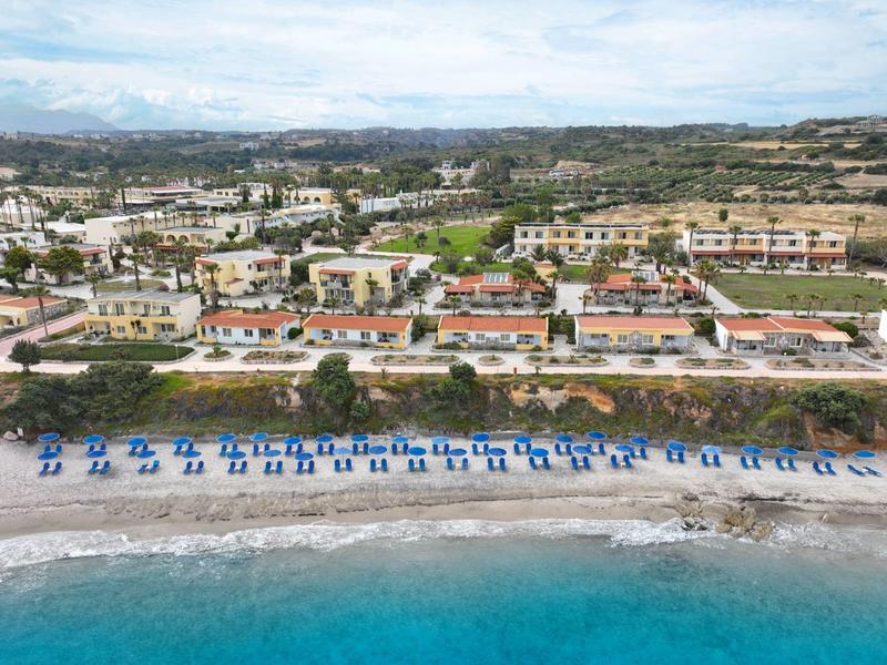 Widok z lotu ptaka na plażę z rzędami niebieskich parasoli i leżaków obok budynków ośrodka.