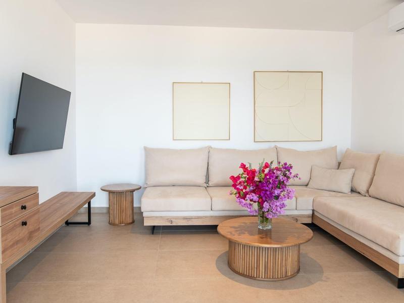 Nowoczesny salon z beżową sofą, drewnianym stolikiem, różowymi kwiatami i telewizorem na ścianie.