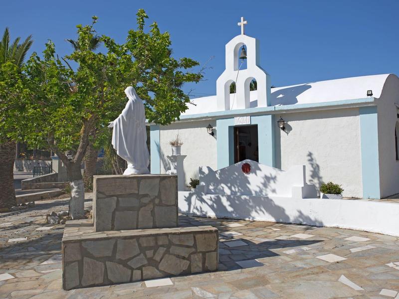 Mała biała kaplica ze statuą na nasłonecznionym dziedzińcu otoczonym drzewami i kamienną nawierzchnią