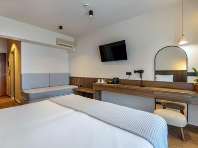Modernes Hotelzimmer mit Bett, Wandfernseher und Schreibtisch mit Spiegel und Hocker.