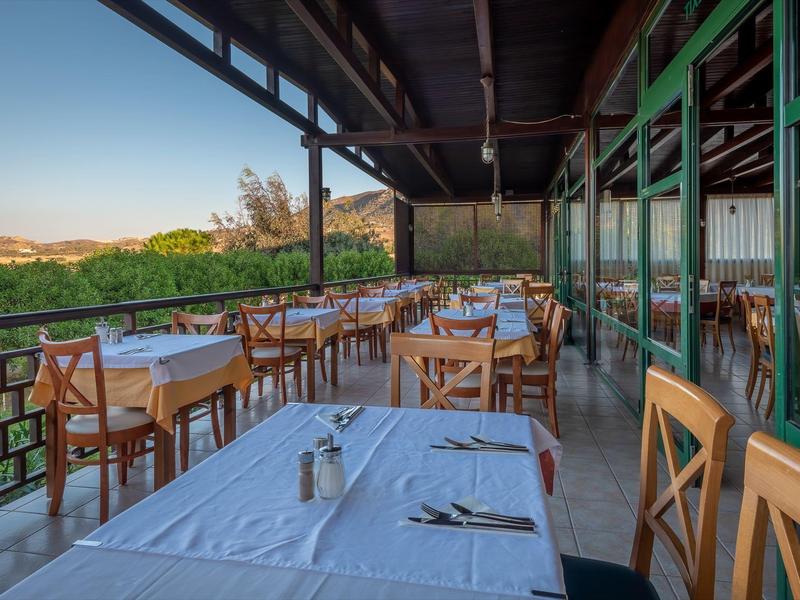 Überdachte Außenterrasse mit Holztischen, weißen Tischdecken und hölzernen Stühlen in warmem Licht.