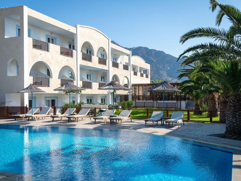 Moderner Hotelkomplex mit großem blauen Pool, Sonnenliegen und Palmen vor Bergkulisse.