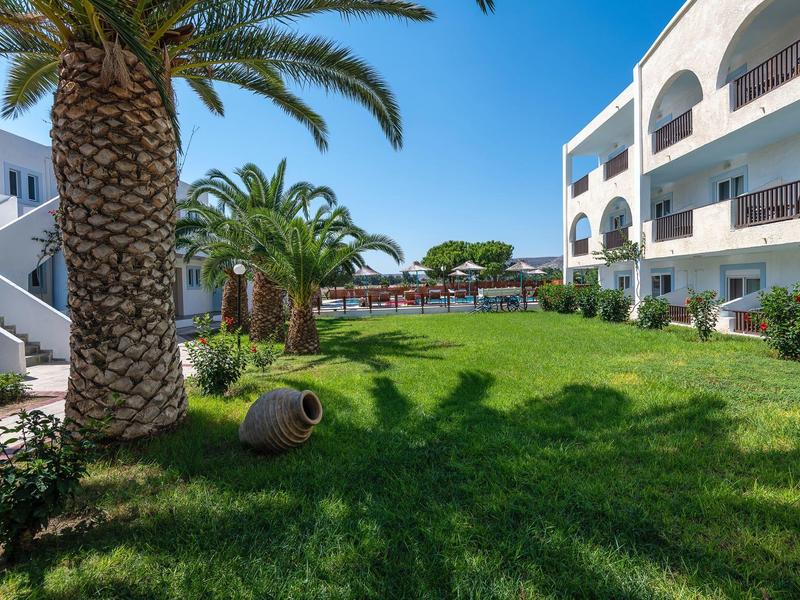 Grüner Garten mit Palmen vor mehrstöckigem weißem Wohnhaus unter blauem Himmel.