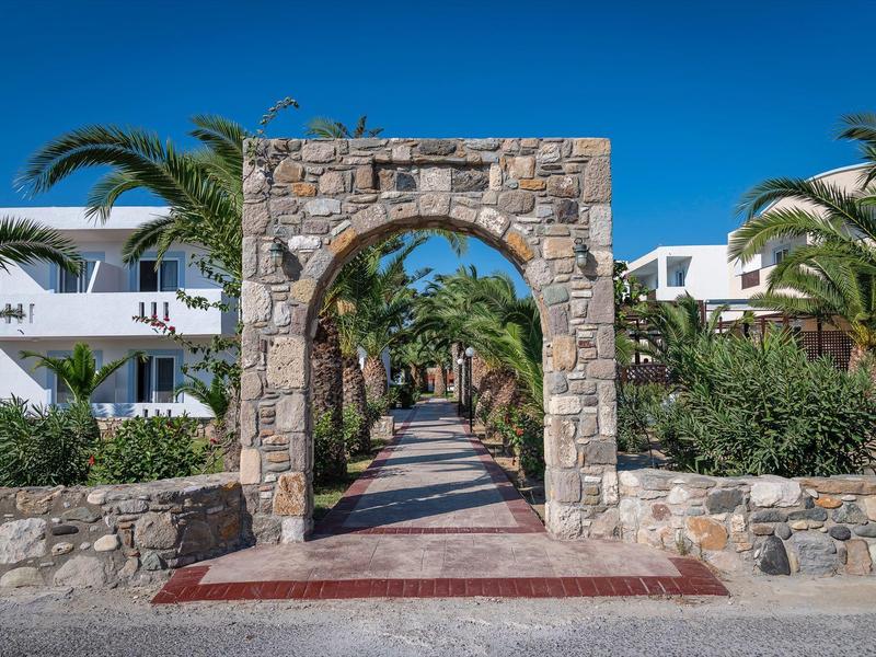 Steinbögen führen zu einem von Palmen gesäumten Weg vor weißen Gebäuden unter klarem blauem Himmel.