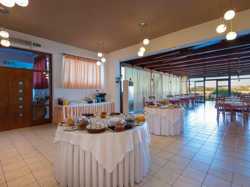 Großer heller Raum mit gedeckten Buffet-Tischen, Stühlen und großen Fenstern, Fliesenboden beige.