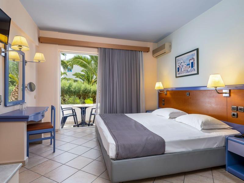 Helles Hotelzimmer mit Doppelbett, Schreibtisch, Balkon und Blick auf grüne Pflanzen.