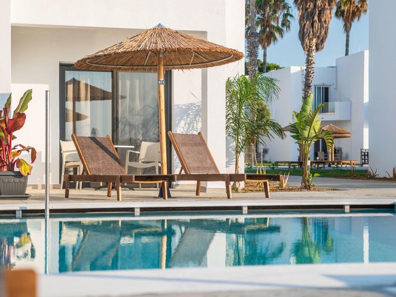 Due sdraio in legno sotto un ombrellone di paglia vicino alla piscina davanti a un edificio di hotel moderno.