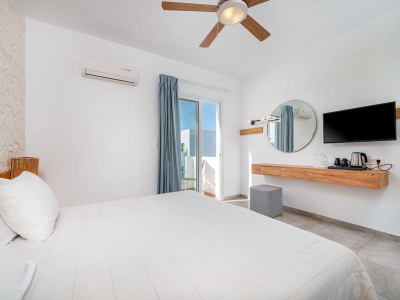 Camera d'albergo luminosa con letto matrimoniale, ventilatore a soffitto, TV e balcone con vista mare.