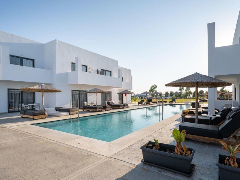 Area piscina esterna moderna con edifici bianchi, lettini e ombrelloni sotto un cielo sereno.