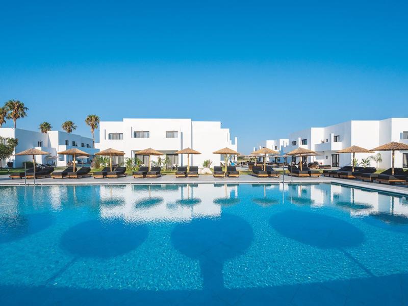 Grande piscina davanti a edifici alberghieri bianchi sotto un cielo azzurro limpido.