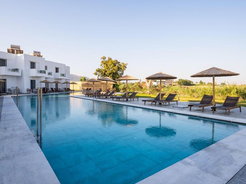 Piscina esterna moderna con lettini e ombrelloni accanto a edifici bianchi sotto un cielo sereno.