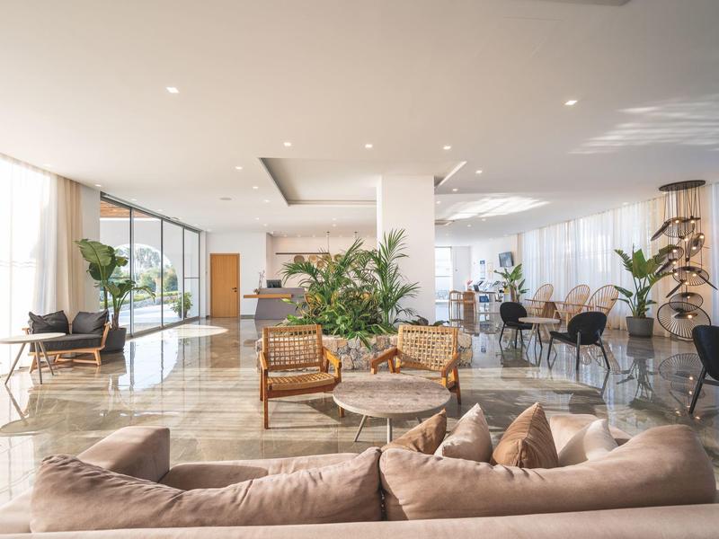 Ampia hall d'hotel moderna e luminosa con aree salotto, piante e grandi finestre.