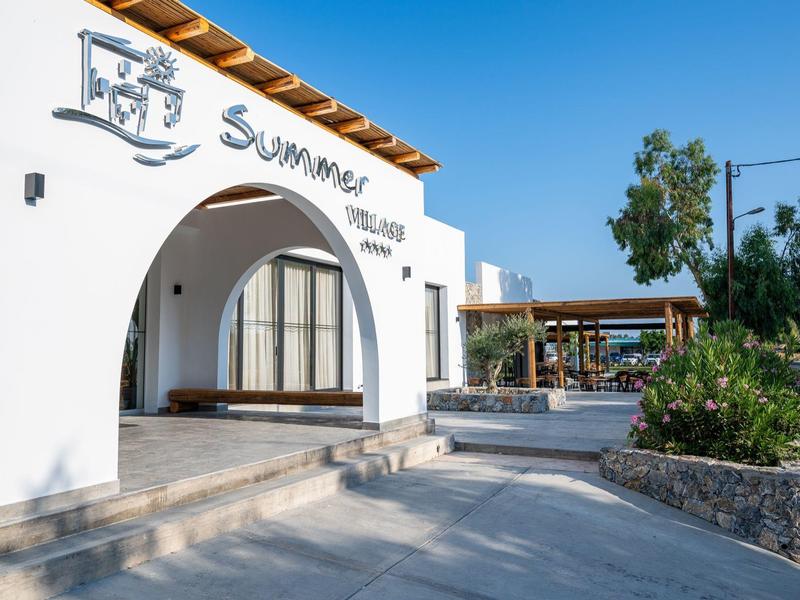 Edificio dell'hotel bianco con arco e cartello 'Summer', area esterna con posti a sedere soleggiata.