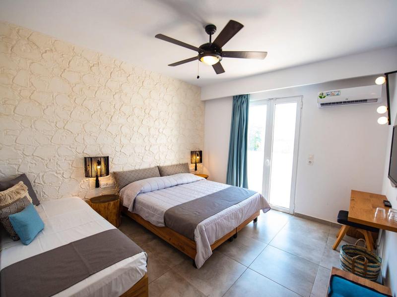 Camera luminosa con letto matrimoniale, letto singolo, ventilatore a soffitto e balcone con tende.