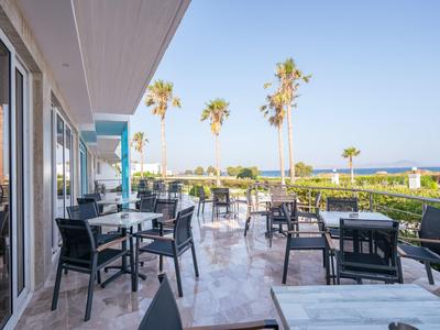 Terrazza con tavoli e sedie davanti a un hotel con vista su palme e mare.