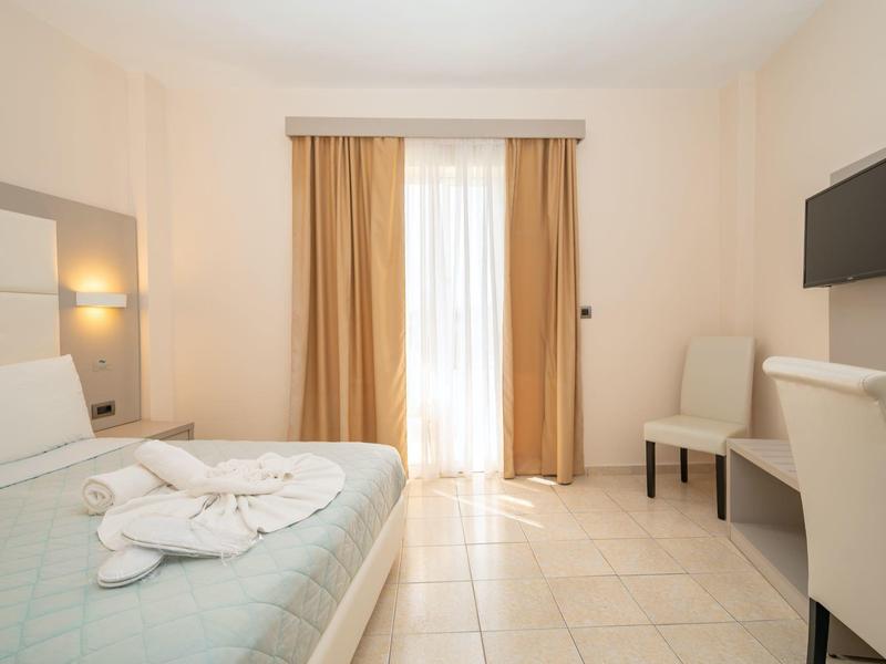 Camera d'hotel luminosa con letto azzurro chiaro, tende marroni e TV a parete.