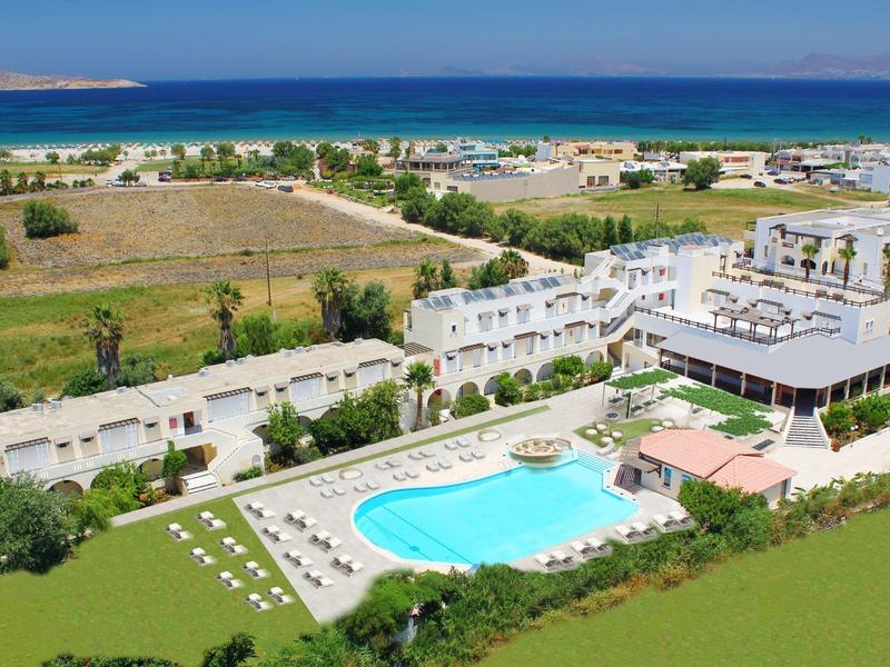 Kompleks hotelowy z dużym basenem, białymi budynkami i morzem w tle.