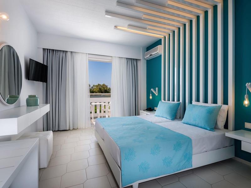 Nowoczesny pokój hotelowy z podwójnym łóżkiem, niebieską ścianą akcentową i balkonem z widokiem na morze.