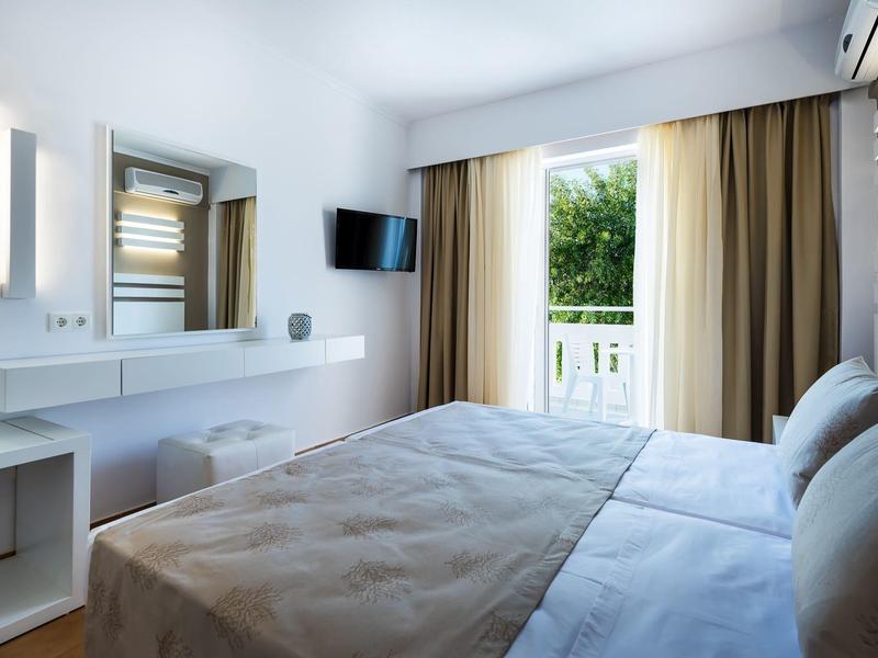 Nowoczesny pokój hotelowy z dużym łóżkiem, biurkiem, telewizorem i balkonem z widokiem na zieleń.