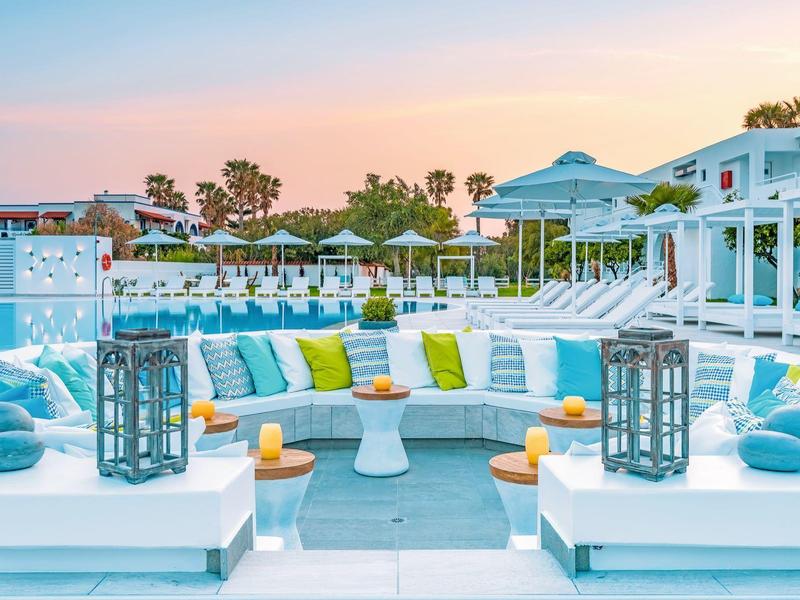 Nowoczesny zewnętrzny salon hotelowy z białymi meblami, kolorowymi poduszkami i palmami w tle.