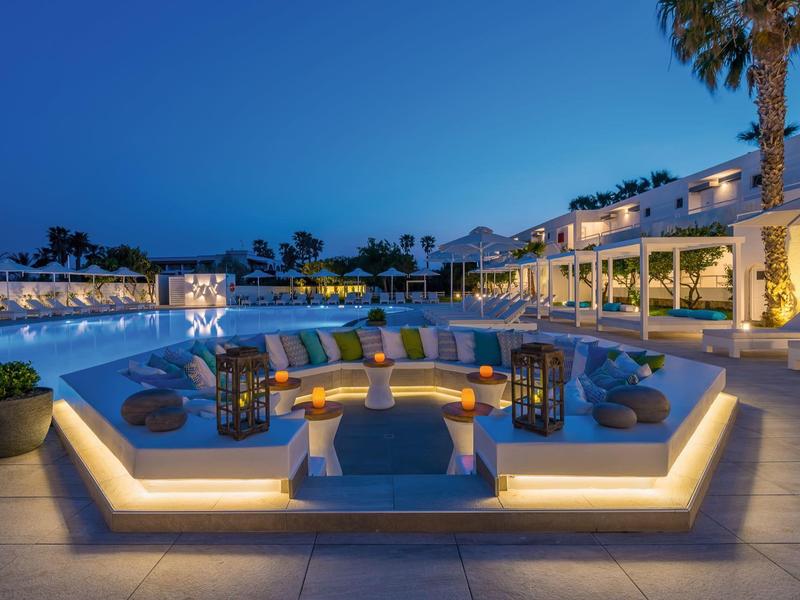 Widok wieczorny na luksusowy bar przy basenie z wygodnymi miejscami do siedzenia i palmami.