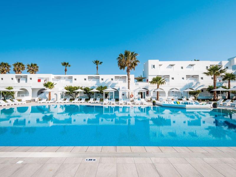 Nowoczesny basen hotelowy z białymi leżakami i palmami pod jasnym niebieskim niebem.