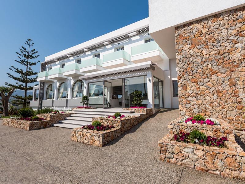 Wejście do nowoczesnego budynku hotelowego z białymi ścianami, szklanymi balustradami i kamiennymi klombami z roślinami.